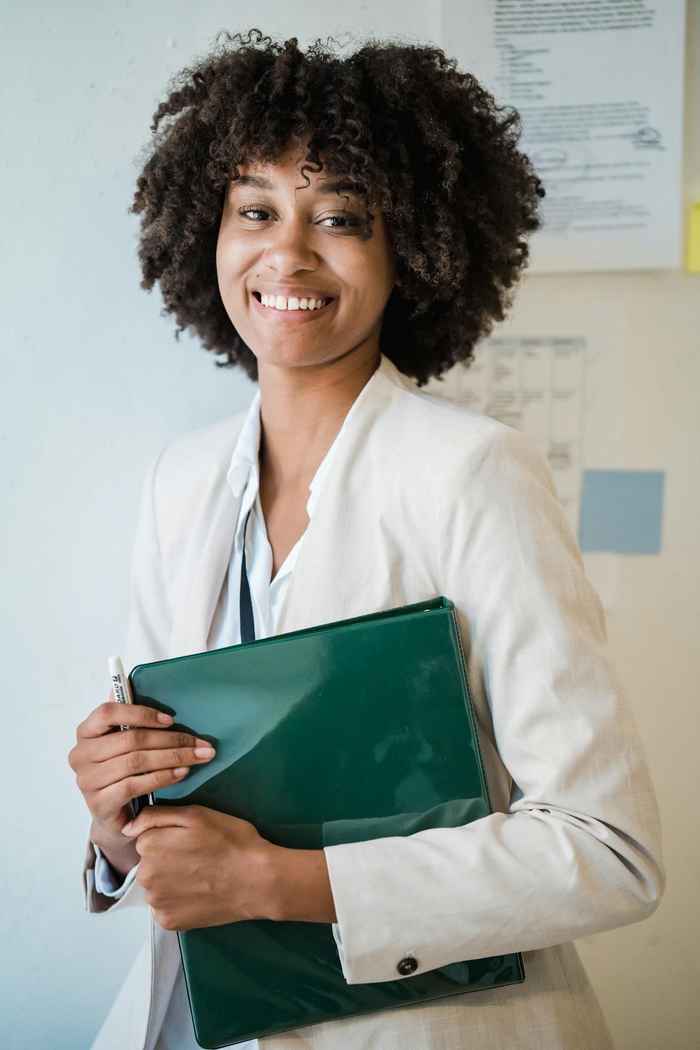 Smiling woman in business attire holding a green file in an office setting.